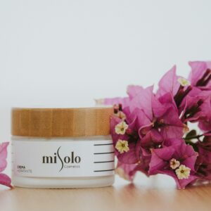 A skincare cream jar with a wooden lid next to vibrant purple bougainvillea flowers on a white background.