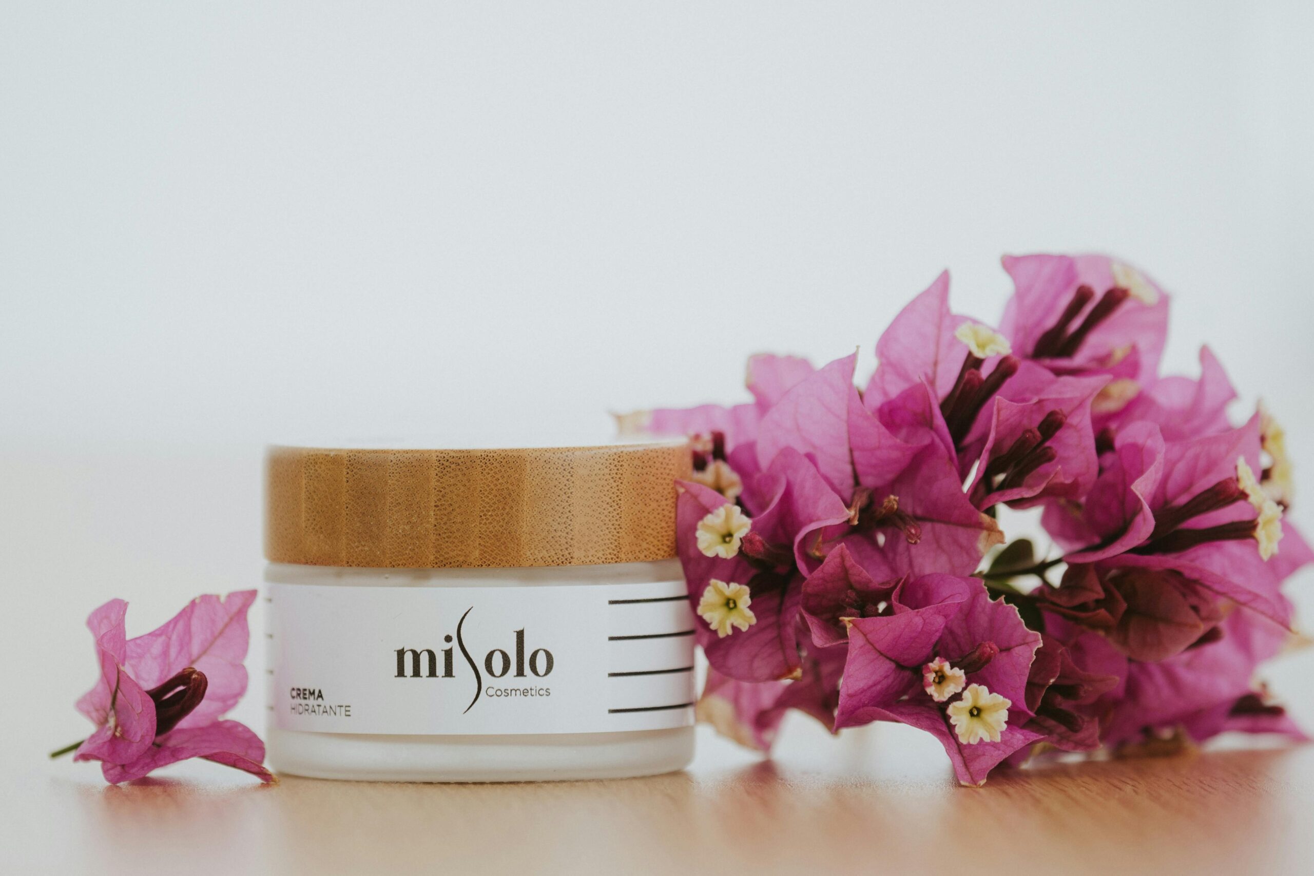 A skincare cream jar with a wooden lid next to vibrant purple bougainvillea flowers on a white background.